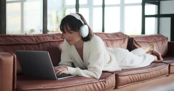 Жінка зручно сидить на дивані, загубившись на екрані ноутбука, одягаючи навушники. Її розслаблена поза та зосереджена перегляд створюють сцену дозвілля та сучасного зв'язку. - Кадри, відео