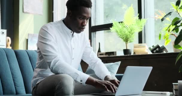 Ein afroamerikanischer Mann sitzt gemütlich auf einem Wohnzimmersofa und tippt eifrig auf seinem Laptop. Sein konzentrierter Ausdruck veranschaulicht die Hingabe an die jeweilige Aufgabe und schafft eine Szene zielgerichteter Arbeit. - Filmmaterial, Video