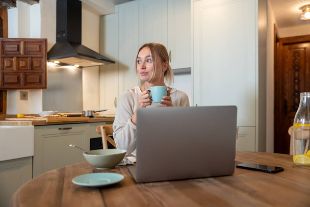Kaukaska kobieta jedząc pyszne śniadanie podczas siedzenia na kuchni i pracy na laptopie  - Zdjęcie, obraz