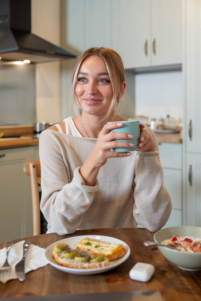 Caucasian female eating delicious healthy breakfast while sitting on kitchen  - Photo, Image