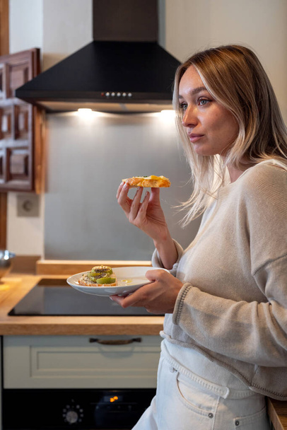 Mujer caucásica en traje casual de pie en el mostrador y comer un poco de pan tostado saludable para el desayuno en la cocina moderna  - Foto, imagen