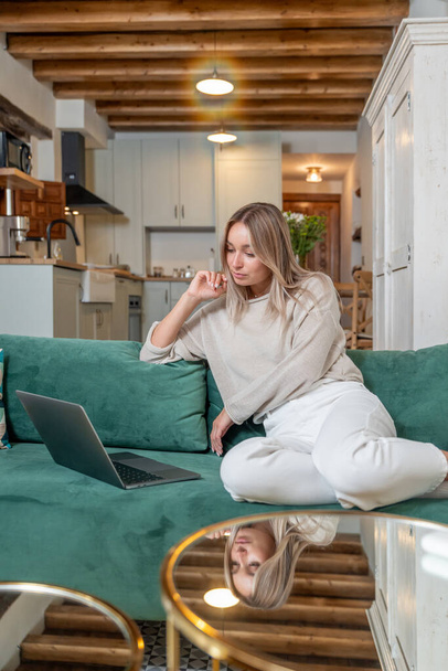 Valkoihoinen nainen istuu sohvalla ja työskentelee etäprojektissa kannettavalla tietokoneella tyylikkäässä olohuoneessa kotona - Valokuva, kuva