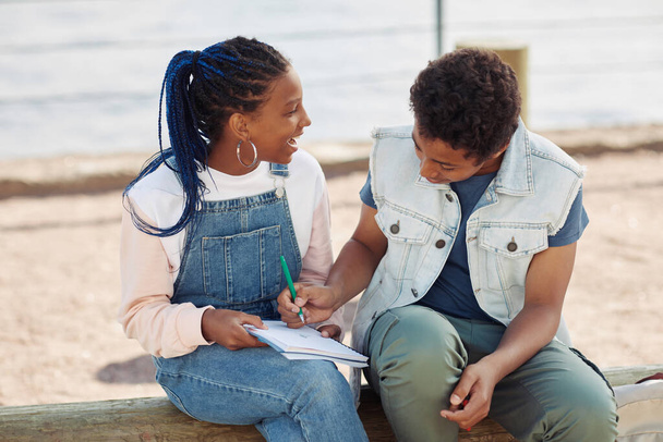Portrait de deux adolescents noirs souriants profitant de cours en plein air et écrivant dans un carnet - Photo, image