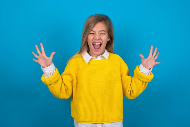 Emotionales kaukasisches Teenie-Mädchen im gelben Pullover über blauer Wand lacht laut, hört lustige Witze oder Geschichten, hebt zufrieden die Handflächen, - Foto, Bild