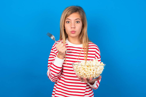 głodny blondynka nastolatek dziewczyna noszenie pasiasty t-shirt nad niebieską ścianą trzymając w ręce widelec nóż chcą smaczne pyszne ciasto pizza - Zdjęcie, obraz