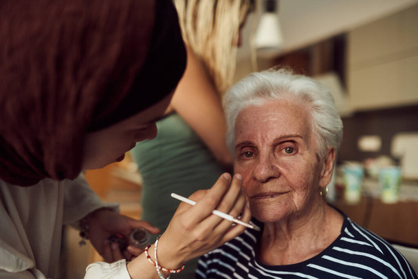 In this heartwarming real-life scene, a girl in a hijab and her sister lovingly apply makeup to their elderly grandmother, preparing her for a special family anniversary celebration, showcasing the - Photo, Image