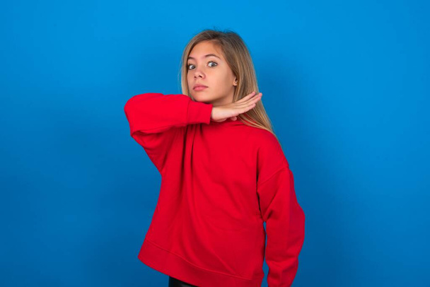bionda teen girl indossa maglione rosso sopra la parete blu tagliando la gola con la mano come coltello, minaccia l'aggressione con violenza furiosa. - Foto, immagini