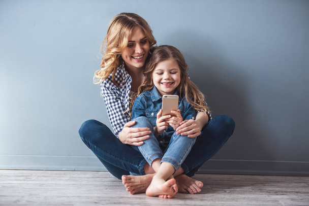 Sevimli küçük kız ve güzel genç annesiyle birlikte yere oturmak, bir akıllı telefon kullanarak ve, gri arka plan üzerinde gülümseyen - Fotoğraf, Görsel