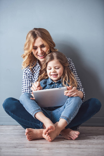 Симпатичная маленькая девочка и ее красивая молодая мама сидят вместе на полу, используя цифровую табличку и улыбаясь на ее фоне
 - Фото, изображение