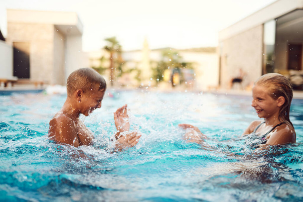 In una villa moderna illuminata dal sole, un fratello e una sorella piccoli spruzzano e giocano gioiosamente in piscina, crogiolandosi nel calore di una perfetta giornata estiva.  - Foto, immagini