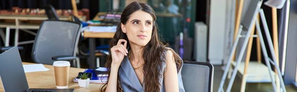pensive businesswoman in casual attire looking away while sitting at laptop and paper cup in office - Foto, Bild