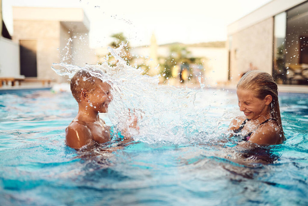 W słonecznej nowoczesnej willi, młodszy brat i siostra radośnie pluskają się i bawią w basenie, pławiąc się w cieple idealnego letniego dnia.  - Zdjęcie, obraz