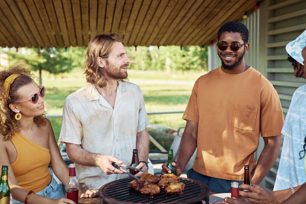 Portrait of two young men grilling meat outdoors with diverse group of friends at barbeque party in Summer - Photo, Image