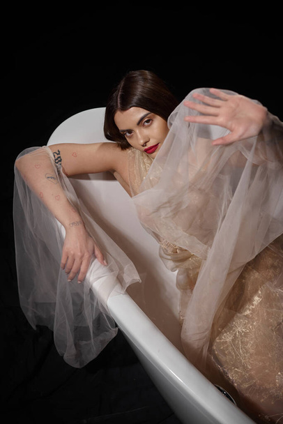 high angle view of graceful model in transparent dress gesturing while lying on bathtub on black - Фото, зображення