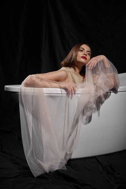 young graceful woman in transparent dress while sitting inside of bathtub on black backdrop - Photo, Image