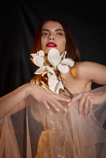 νεαρή ονειρεμένη γυναίκα με ρομαντικό διάφανο φόρεμα που κρατά λευκό ανθισμένο λουλούδι σε μαύρο φόντο - Φωτογραφία, εικόνα