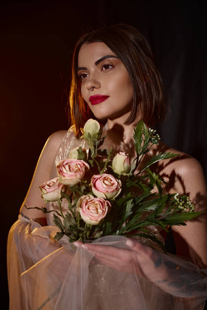 charmante jonge vrouw in romantische transparante jurk houden bloeiende rozen op zwarte achtergrond - Foto, afbeelding