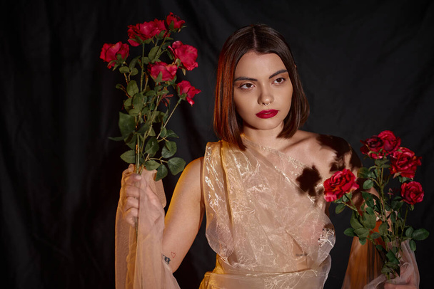 Romantik, şeffaf elbiseli genç bir kadın elinde çiçek açan kırmızı güller tutuyor. - Fotoğraf, Görsel