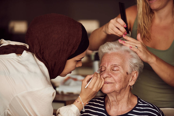 Ebben a szívmelengető valós jelenetben, egy hidzsábos lány és a nővére szeretettel sminkelik ki idős nagymamájukat, felkészítve őt egy különleges családi évfordulós ünnepségre, bemutatva a - Fotó, kép