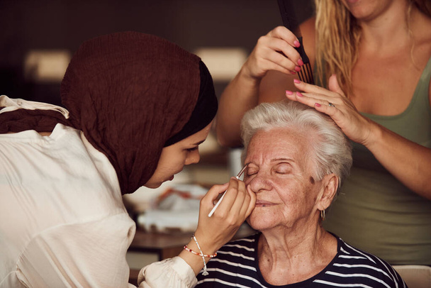 W tej serdecznej scenie prawdziwego życia, dziewczyna w hidżabie i jej siostra z miłością nakładają makijaż na starszą babcię, przygotowując ją do specjalnej uroczystości rocznicowej rodziny, pokazując - Zdjęcie, obraz