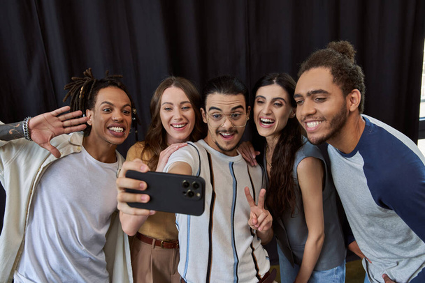 Gözlüklü neşeli adam zafer işareti gösteriyor ve çoklu etnik gruplarla akıllı telefondan selfie çekiyor. - Fotoğraf, Görsel