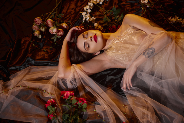 charming tattooed young woman in romantic transparent dress lying among blooming flowers - Photo, Image
