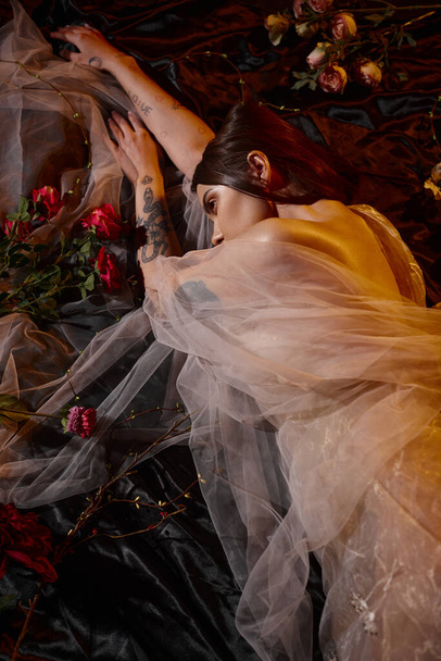 charming and tattooed young model in romantic transparent dress lying among blooming flowers - 写真・画像