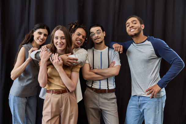 freudige multiethnische Kollegen umarmen und blicken in die Kamera vor schwarzem Hintergrund in modernen Büros - Foto, Bild