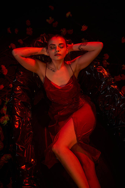 felső nézet, bájos fiatal nő piros slip ruha feküdt fekete fürdőkádban között gyönyörű virágok - Fotó, kép