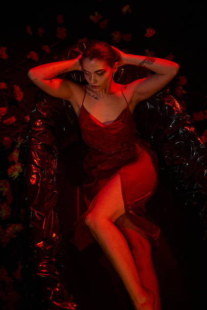 κόκκινο φως, αισθησιακό νεαρή γυναίκα σε ολίσθηση φόρεμα που βρίσκεται σε μαύρη μπανιέρα ανάμεσα σε όμορφα λουλούδια - Φωτογραφία, εικόνα