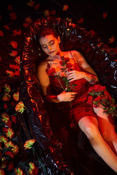 jovem bonita em vestido de deslizamento molhado deitado na banheira preta com flores florescendo, luz vermelha - Foto, Imagem
