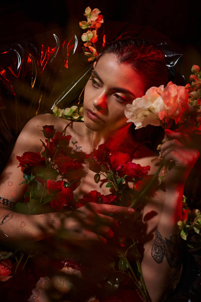 álmodozó fiatal nő nedves piros ruhában fekve fekete fürdőkádban virágzó virágok között - Fotó, kép