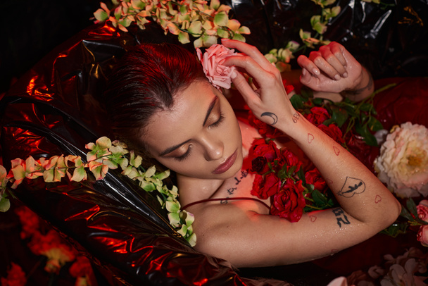 attractive young woman in wet red dress lying in black bathtub among blooming flowers, top view - Photo, Image