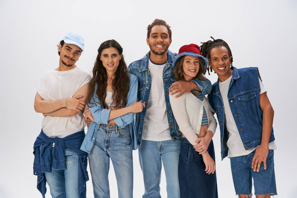 веселые межрасовые друзья в модной и джинсовой одежде, обнимающие на сером фоне, уличная мода - Фото, изображение