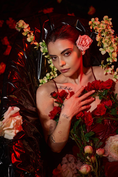 tattooed woman in wet red dress looking at camera and lying in bathtub among blooming flowers - 写真・画像
