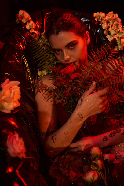 красный свет на лице привлекательной влажной женщины с пирсингом и татуировкой, лежащей среди различных цветов - Фото, изображение