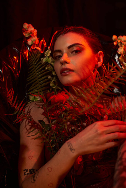 κόκκινο φως στο πρόσωπο της όμορφης νεαρής γυναίκας με σκουλαρίκια και τατουάζ που βρίσκεται ανάμεσα σε διάφορα λουλούδια - Φωτογραφία, εικόνα