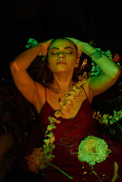 luz verde, mujer joven atractiva con el pelo mojado posando con los ojos cerrados entre flores florecientes - Foto, Imagen