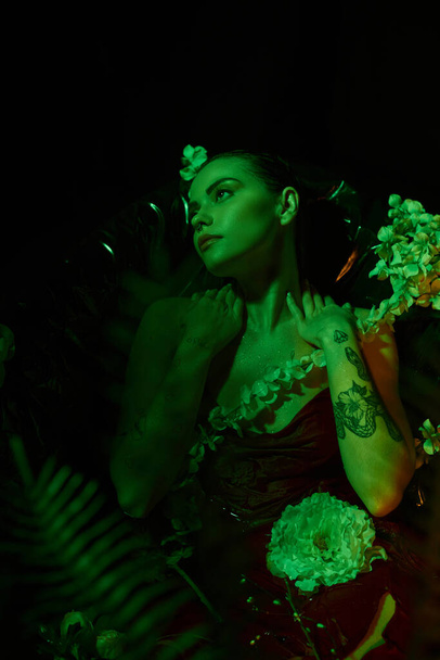 зелене світло, мрійлива молода жінка з мокрим волоссям дивиться в сторону і позує серед квітів, елегантність - Фото, зображення