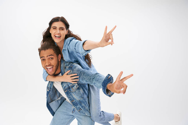 overjoyed couple in trendy denim wear showing victory signs while having fun on grey, piggybacking - Photo, Image