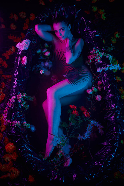 θηλυκή ομορφιά, νεαρή γυναίκα ξαπλωμένη ανάμεσα σε φύλλα φοίνικα και λουλούδια στην μπανιέρα, μπλε και μοβ φώτα - Φωτογραφία, εικόνα