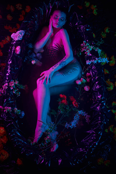 θηλυκή ομορφιά, όμορφη γυναίκα ξαπλωμένη ανάμεσα σε φύλλα φοίνικα και λουλούδια στην μπανιέρα, μπλε και μοβ φώτα - Φωτογραφία, εικόνα
