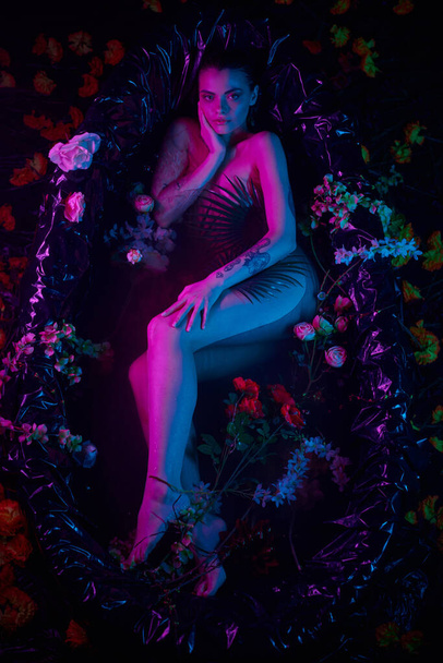 θηλυκή χάρη, όμορφη γυναίκα ξαπλωμένη ανάμεσα σε φύλλα φοίνικα και λουλούδια στην μπανιέρα, μπλε και μοβ φώτα - Φωτογραφία, εικόνα