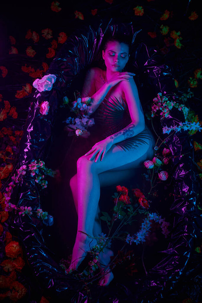 tiro de moda, mujer joven acostada entre hojas de palma y flores en bañera, luces azules y púrpuras - Foto, imagen