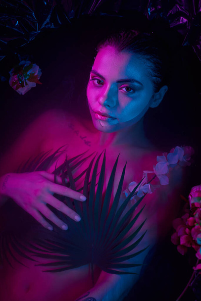 fashion shot, charming woman lying among palm leaves and flowers in water, blue and purple lights - Photo, Image