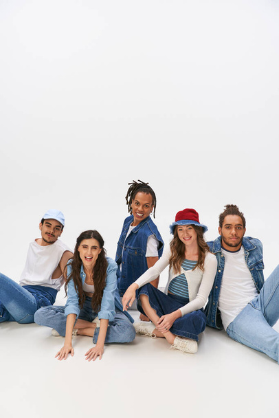 šťastný multietnické přátelé v módní a džínové oblečení sedí a dívá se na kameru na šedém pozadí - Fotografie, Obrázek