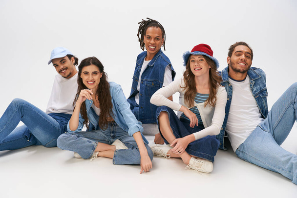 stylish multiethnic friends in casual street wear sitting and smiling at camera on grey backdrop - Photo, Image