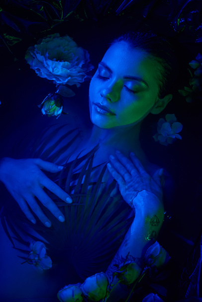 vrouwelijke schoonheid, mooie jonge vrouw liggend tussen palmbladeren en bloemen in water, blauw licht - Foto, afbeelding