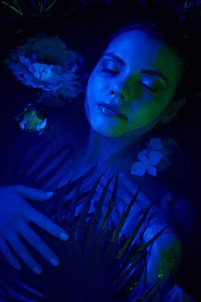 женская красота, расслабленная молодая женщина лежит среди пальмовых листьев и цветов в воде, голубой свет - Фото, изображение