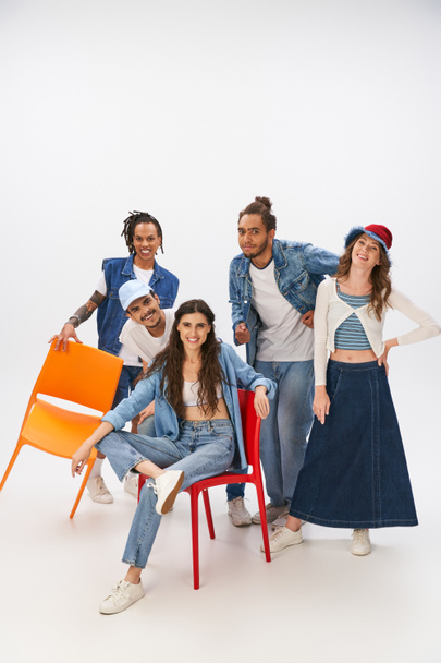 smiling woman sitting on chair near multiethnic friends in trendy casual attire on grey backdrop - Photo, Image